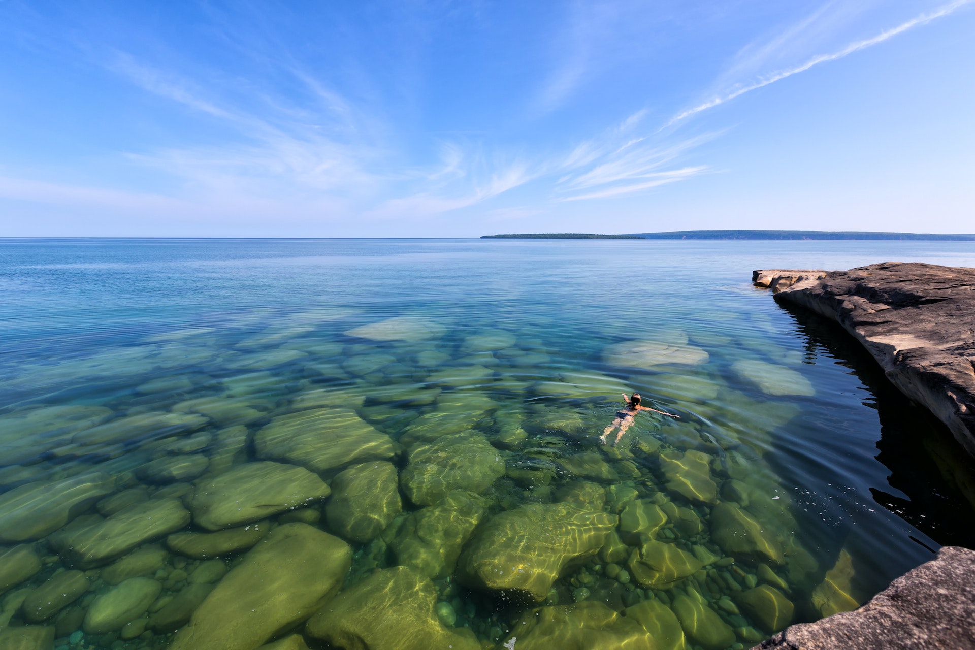 Девушка плавает в озере Верхнее на верхнем полуострове озера Мичиган.  Сквозь стекло видны скалы, похожие на чистейшую воду.  На фото вдалеке находится Национальный берег озера Рокс.